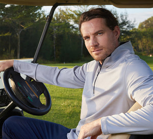 A man sitting in a golf cart on a golf course, wearing a light gray Pinstripe Quarter Zip Pullover and navy blue pants, looking relaxed and confident.