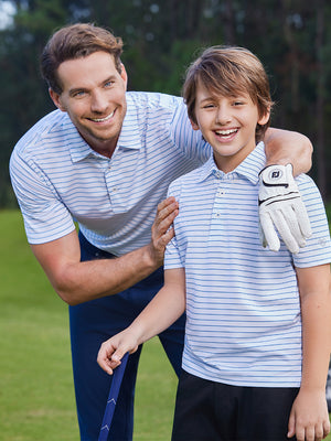 Father resting hand on son's shoulder on golf course, both wearing youth performance golf polos in white/light blue stripe